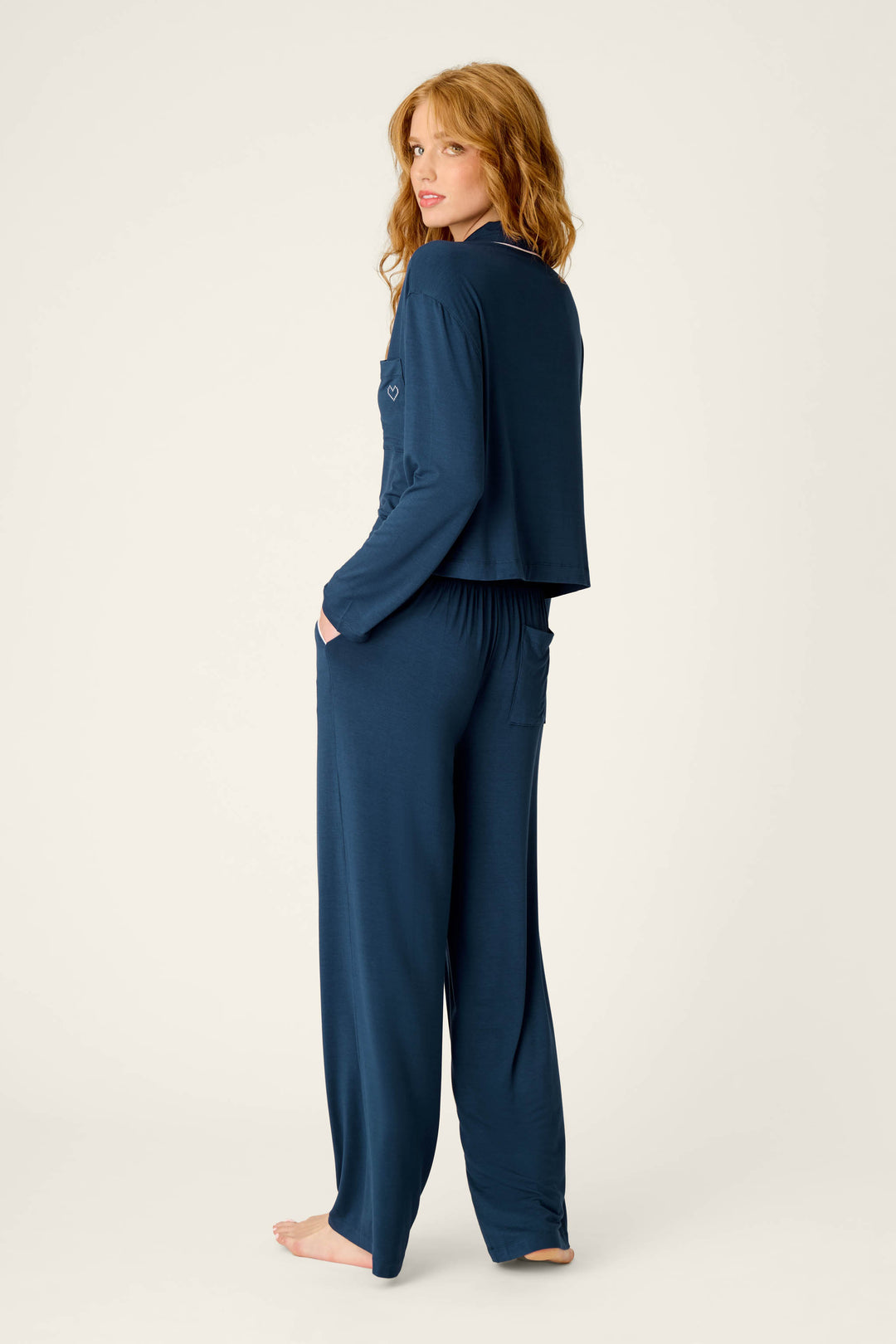 A woman with wavy red hair, standing barefoot against a plain light background and looking over her shoulder, is wearing the Soiree Dreams Deep Navy Long Sleeve PJ Set by P.J. Salvage.