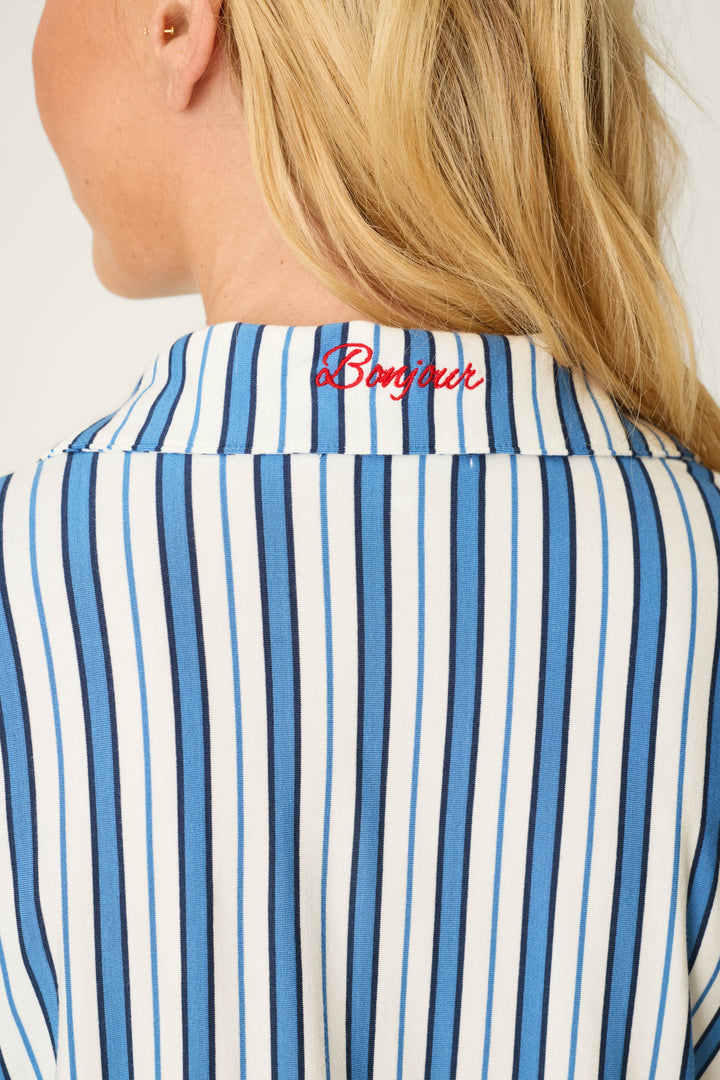 A woman with long blonde hair wears the Soiree Dreams Blue Stripe Long Sleeve & Shorts PJ Set by P.J. Salvage, featuring a white shirt with vertical blue stripes and red "Bonjour" embroidery on the collar at the back of her neck.