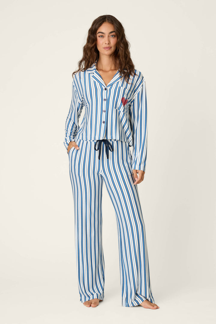 A woman stands barefoot against a plain light background, wearing the P.J. Salvage Soiree Dreams Blue Stripe Long Sleeve PJ Set featuring blue and white vertical stripes, a button-up long sleeve top, matching drawstring pants, and hands in her pockets.