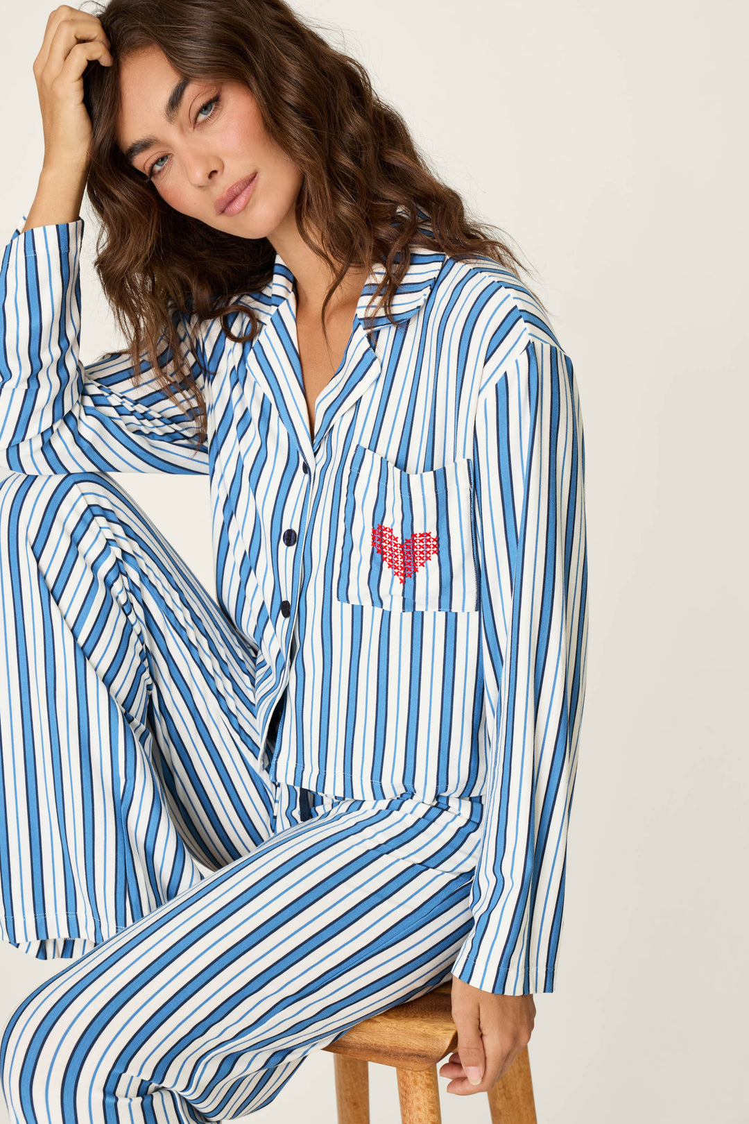 A woman sits on a stool, looking relaxed with one hand touching her head, wearing the P.J. Salvage Soiree Dreams Blue Stripe Long Sleeve PJ Set featuring blue and white stripes and a red heart on the chest pocket.