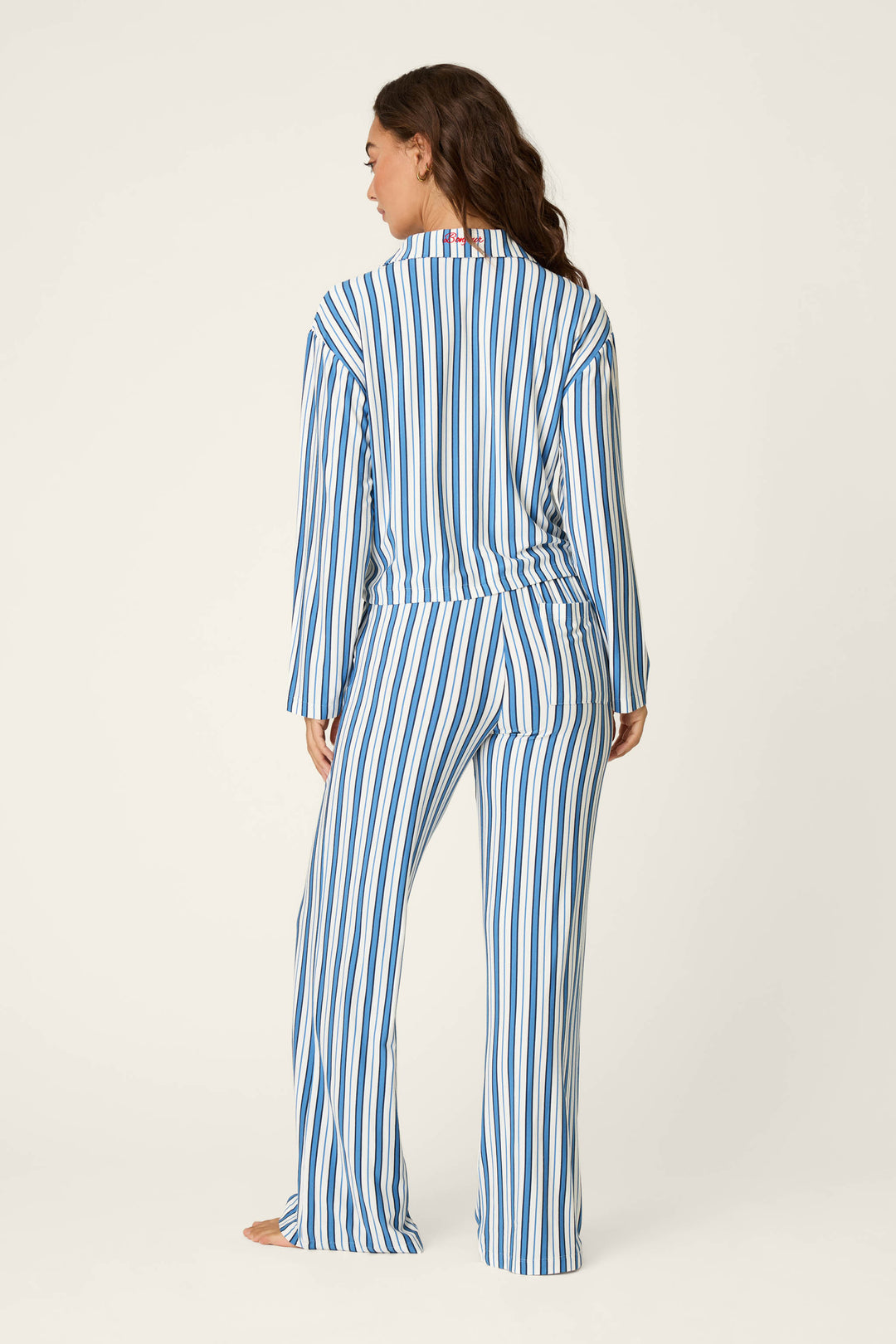 A woman stands barefoot, facing away, wearing the P.J. Salvage Soiree Dreams Blue Stripe Long Sleeve PJ Set—featuring blue and white vertical stripes on both the top and matching pants—against a plain light background.