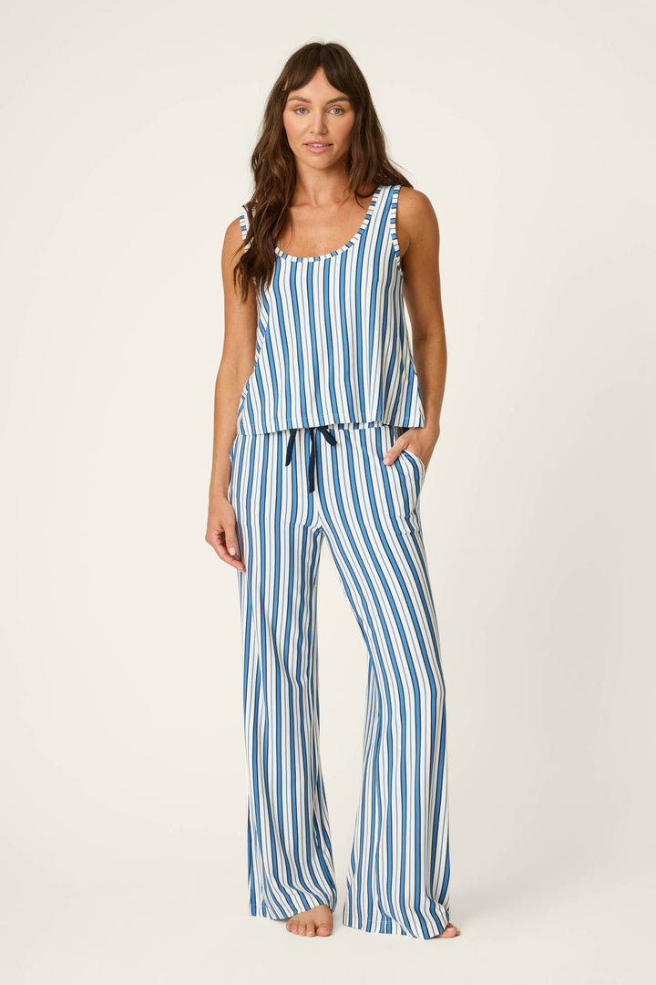 A woman stands barefoot against a plain light background, wearing the P.J. Salvage Soiree Dreams Blue Stripe Tank & Pants PJ Set featuring sleeveless styling, blue-and-white vertical stripes, loose-fitting pants, and a comfortable drawstring waist.