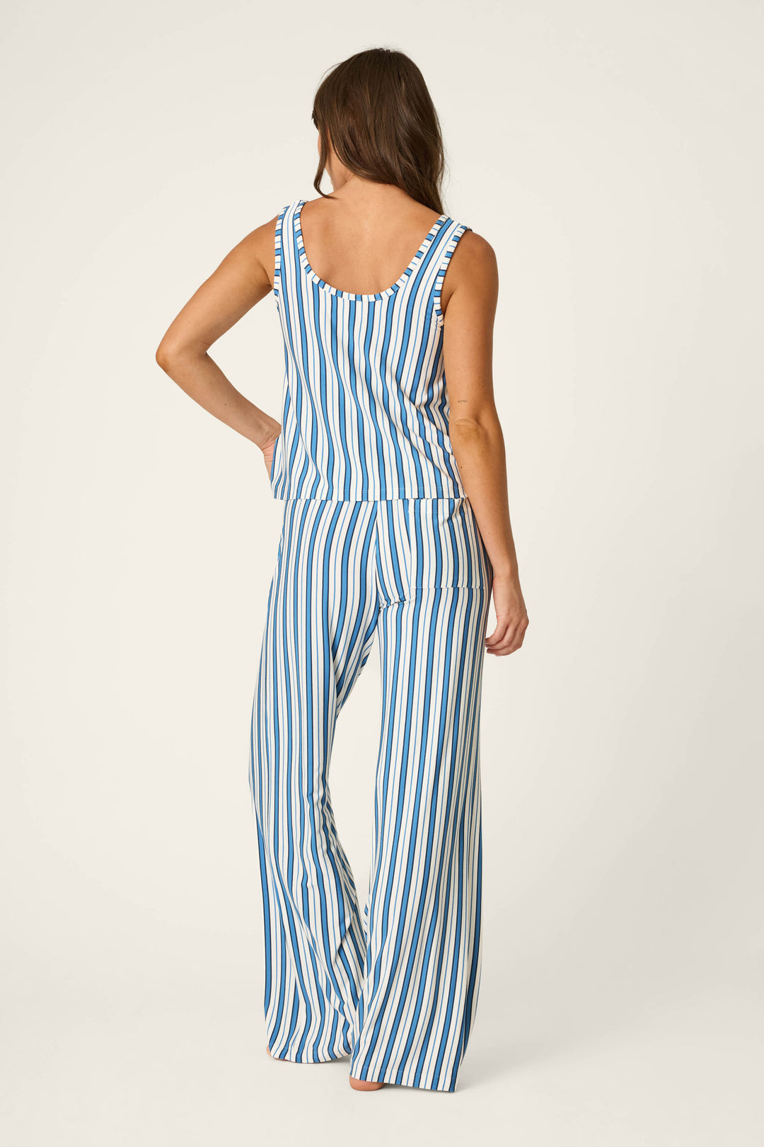 A woman stands with her back to the camera, wearing the P.J. Salvage Soiree Dreams Blue Stripe Tank & Pants PJ Set—featuring a sleeveless top and wide-leg pants with vertical blue and white stripes—against a plain, light-colored background.