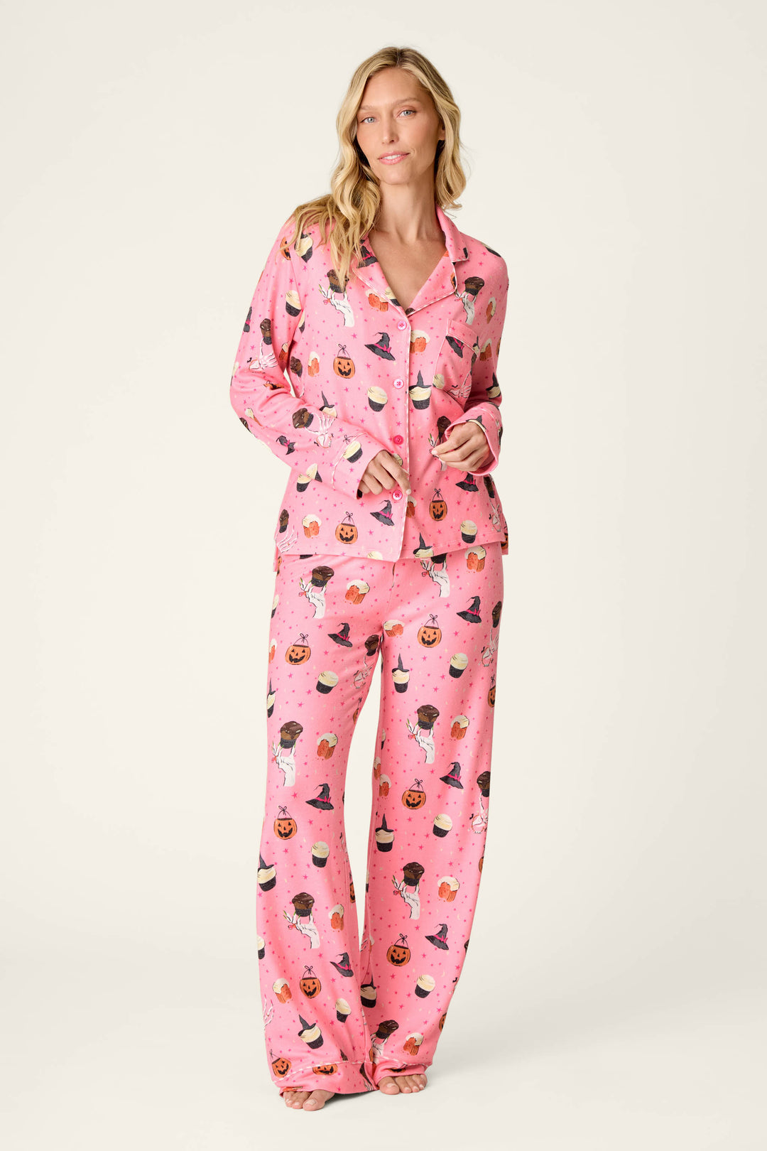 A woman stands barefoot against a plain light background wearing the Sweet and Spooky Halloween Print Long Sleeve PJ Set by P.J. Salvage—a pink pajama set adorned with a colorful Halloween-themed print featuring witches, pumpkins, and black cats.