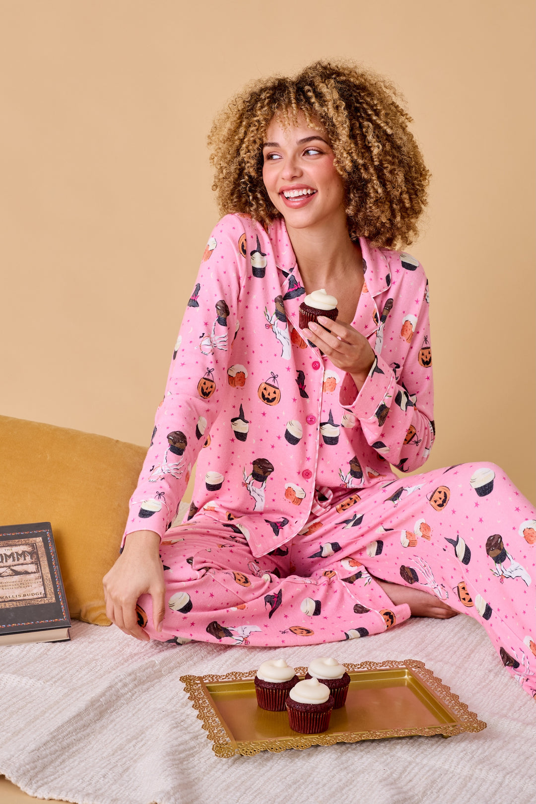 A woman with curly hair wearing a pink long-sleeve pajama set with a spooky cupcake & jack o lantern print, sitting on a bed and holding a cupcake. A gold tray with more cupcakes and a book are in the foreground, against a warm beige background.