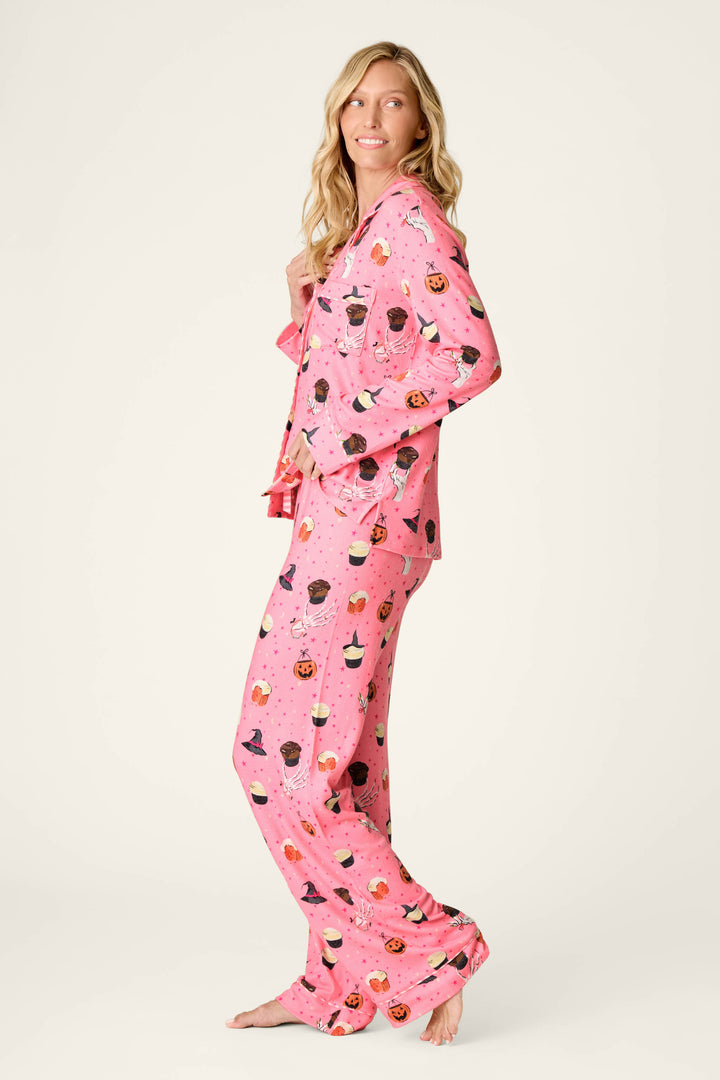 A woman with long blonde hair stands barefoot and smiling against a neutral off-white background, wearing the P.J. Salvage Sweet and Spooky Halloween Print Long Sleeve PJ Set featuring playful dessert-inspired patterns in pink hues.