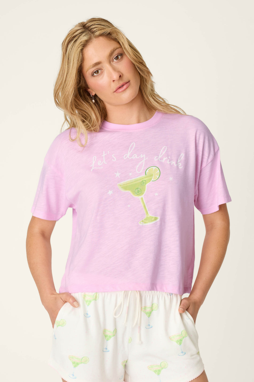 A blonde woman wears the Softee "Let's Day Drink" Graphic Print T-Shirt by P.J. Salvage, paired with white shorts featuring small margarita prints, standing against a plain light background.