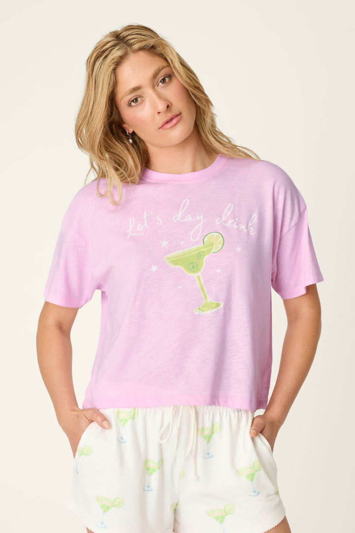 A blonde woman wears the Softee "Let's Day Drink" Graphic Print T-Shirt by P.J. Salvage, paired with white shorts featuring small margarita prints, standing against a plain light background.