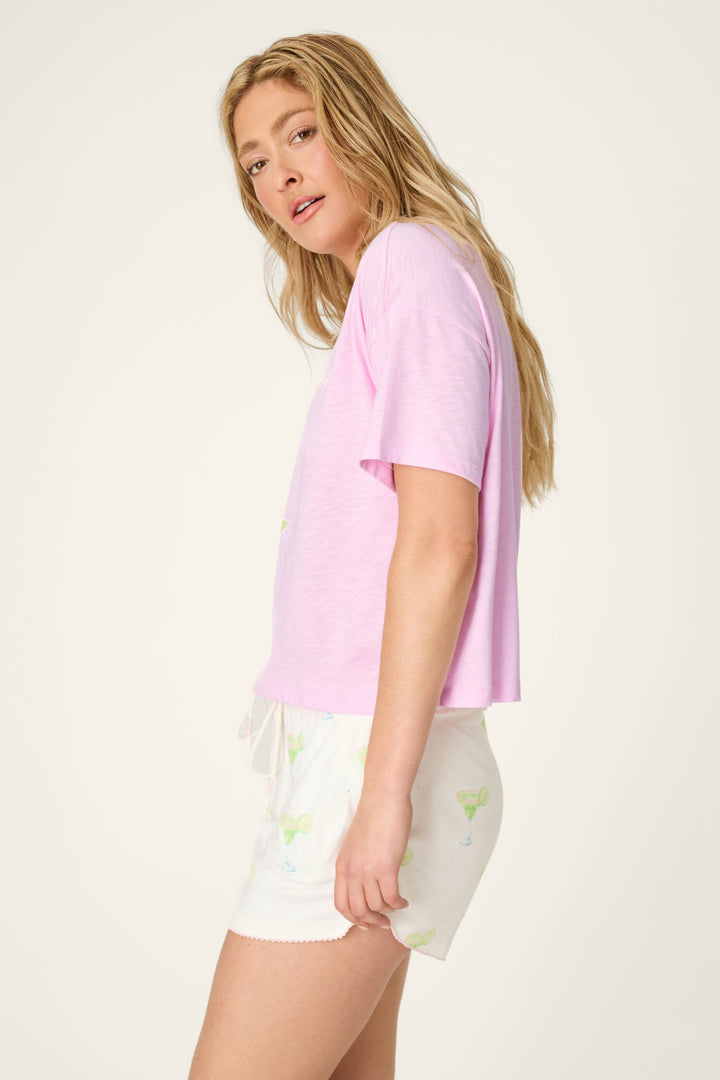A young woman with long blonde hair wears the P.J. Salvage Softee "Let's Day Drink" Graphic Print T-Shirt and white shorts with a subtle green pattern, standing sideways and looking at the camera against a plain background.