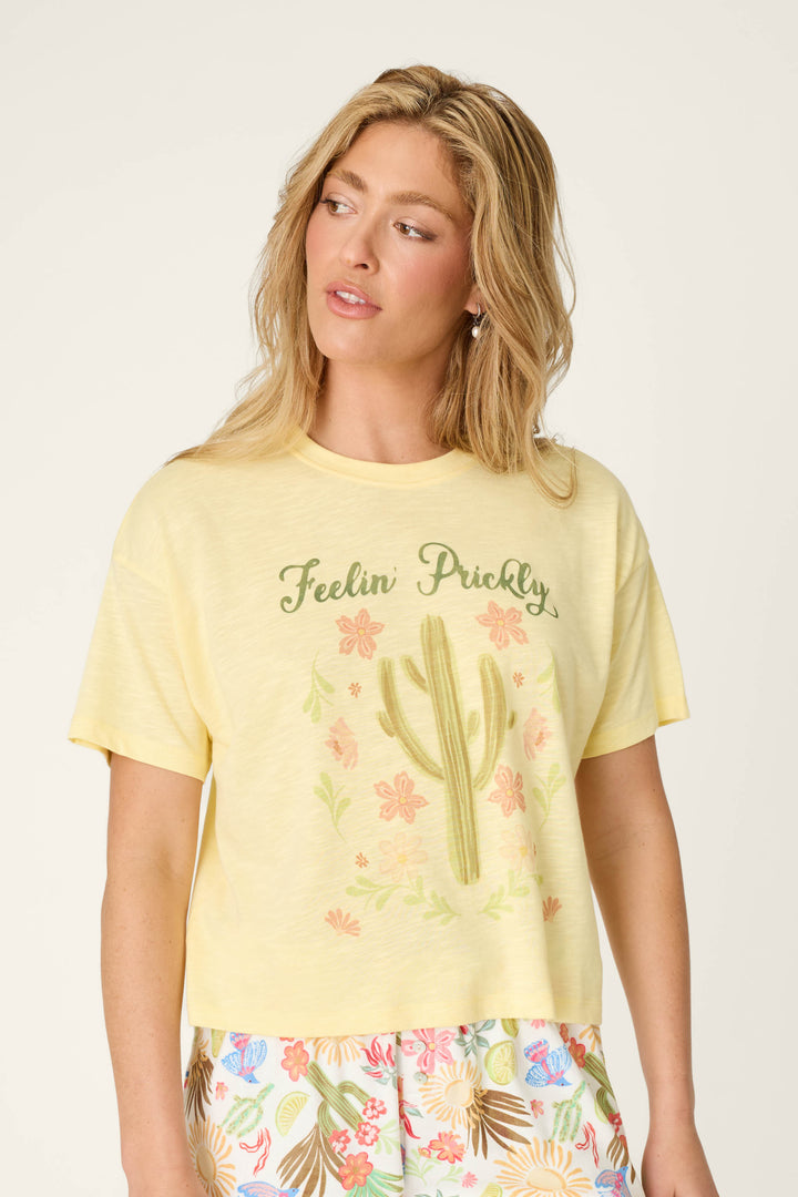A woman with long blonde hair wears the P.J. Salvage Softee "Feelin' Prickly" Graphic Print T-Shirt with a cactus and flower design, paired with colorful leaf-patterned shorts, as she looks off to the side.