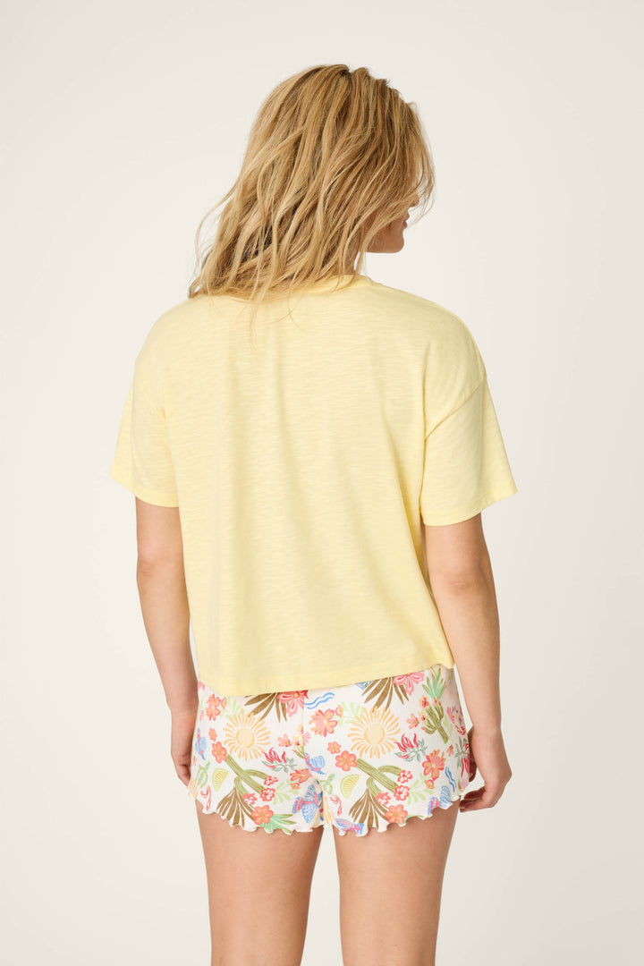 A woman with blonde hair, seen from behind, wears the P.J. Salvage Softee "Feelin' Prickly" Graphic Print T-Shirt in pale yellow and colorful floral shorts against a light background.