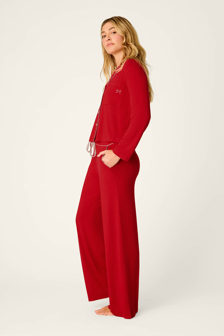 A woman with long blonde hair stands barefoot in profile against a plain white background, wearing the P.J. Salvage Sweetheart Dreams Red Long Sleeve PJ Set, featuring a loose fit with wide-leg pants and a button-up top.