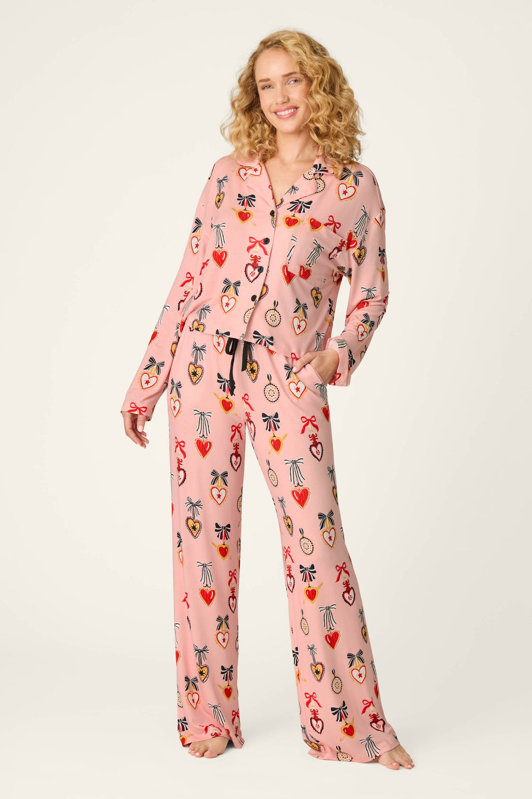 A woman with curly blonde hair stands barefoot against a plain light background, smiling with one hand in her pocket while wearing the P.J. Salvage Sweetheart Dreams Love Lockets Print Long Sleeve PJ Set featuring colorful heart and ribbon designs.