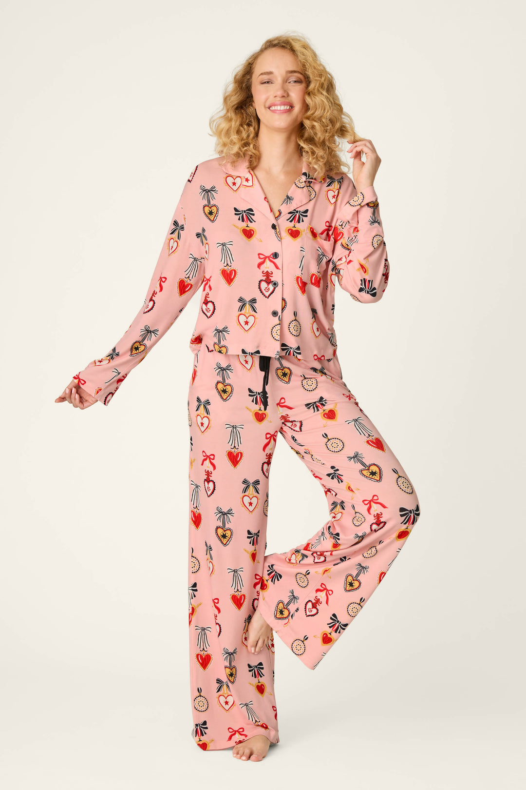 A woman with curly blonde hair wears the P.J. Salvage Sweetheart Dreams Love Lockets Print Long Sleeve PJ Set, standing on one leg and smiling against a plain background.