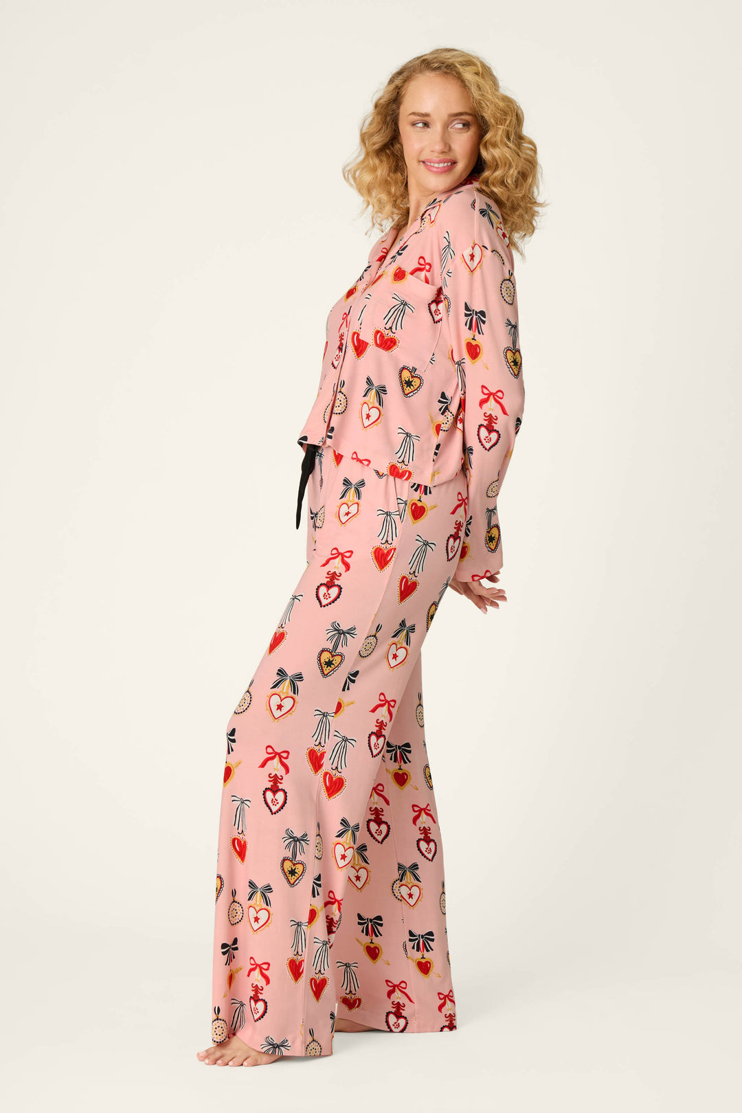 A woman with wavy blonde hair stands barefoot against a plain light background, smiling sideways while wearing the P.J. Salvage “Sweetheart Dreams Love Lockets Print” Long Sleeve PJ Set, featuring matching pink pajamas adorned with colorful heart and bow patterns.