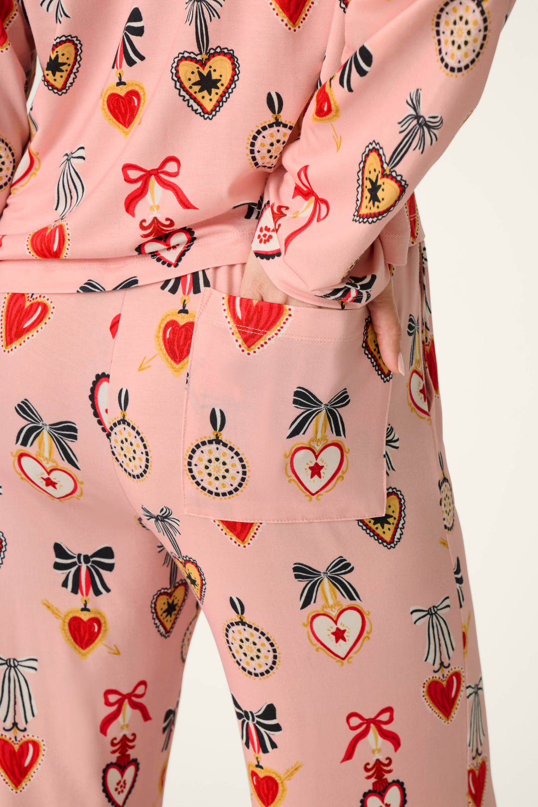 A person is wearing the P.J. Salvage Sweetheart Dreams Love Lockets Print Long Sleeve PJ Set, featuring red and yellow heart and bow motifs. They have one hand placed in the pocket of the pants, standing against a plain, off-white background.