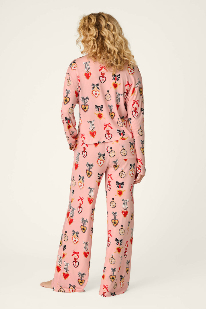 A person with wavy blonde hair, standing barefoot and facing away from the camera against a plain background, wears the P.J. Salvage Sweetheart Dreams Love Lockets Print Long Sleeve PJ Set—loose-fitting pink pajamas adorned with colorful hearts, crowns, and bows.