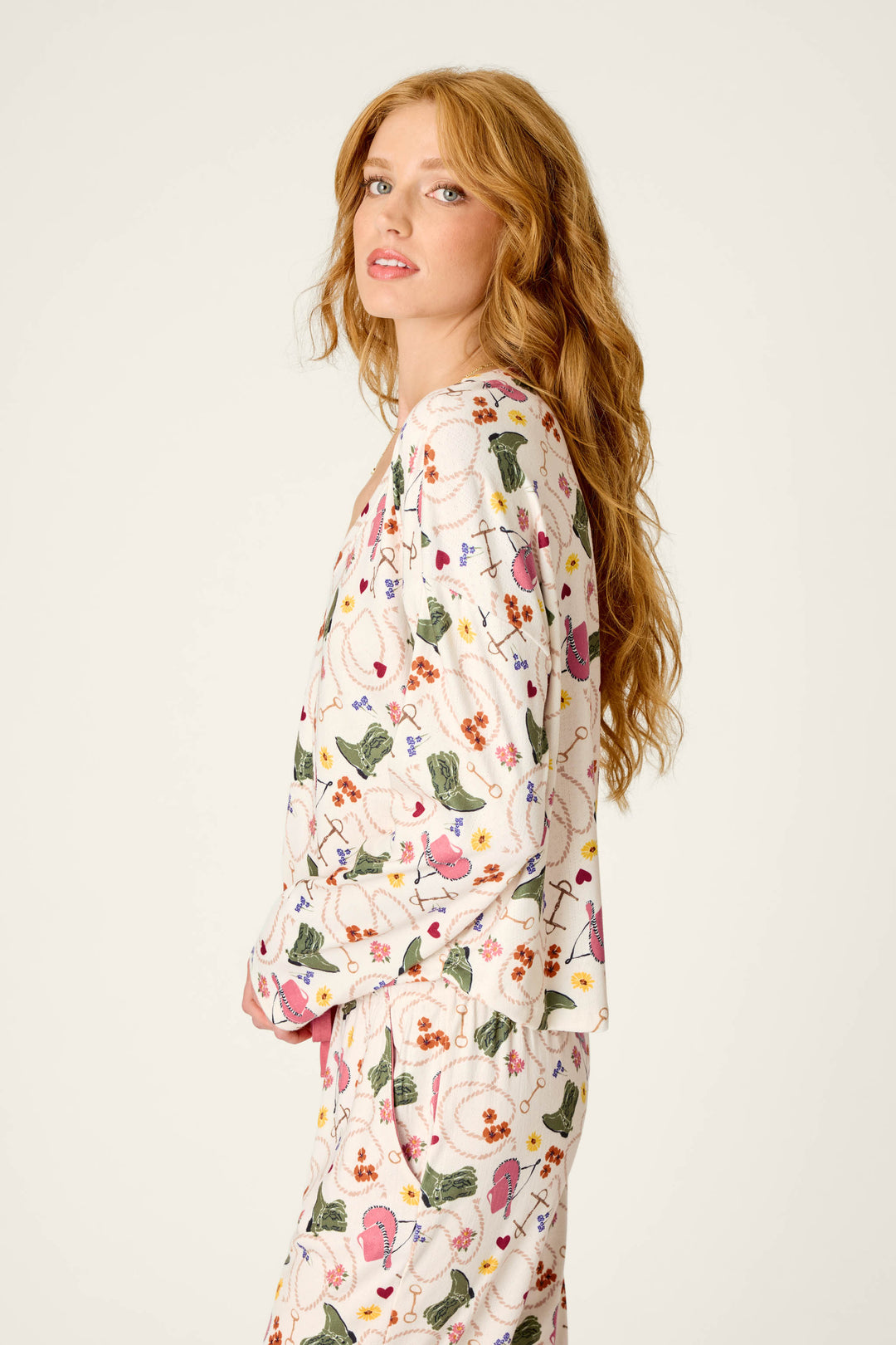 A woman with long wavy red hair, wearing the P.J. Salvage Saddle Up Western Print Long Sleeve V-Neck Top, stands sideways against a plain light background.