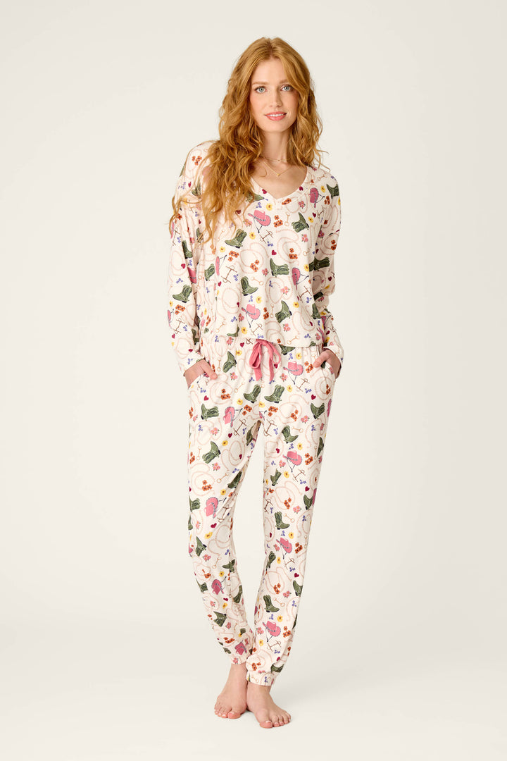 A woman with long, wavy hair stands barefoot, smiling slightly with her hands in her pockets. She is wearing the P.J. Salvage Saddle Up Western Print Long Sleeve V-Neck Top.