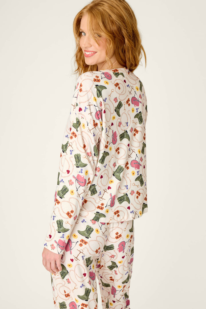 A woman with red hair smiles while looking back over her shoulder. She is wearing the P.J. Salvage Saddle Up Western Print Long Sleeve V-Neck Top, which features playful patterns and images on a white background.