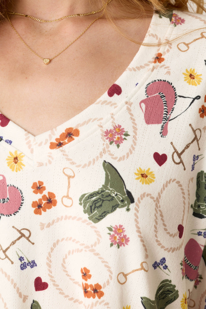 A close-up of the P.J. Salvage Saddle Up Western Print Long Sleeve V-Neck Top, featuring a pattern with flowers, hearts, horse bits, riding gloves, and ropes. A woman wearing a small gold heart necklace is partially visible.