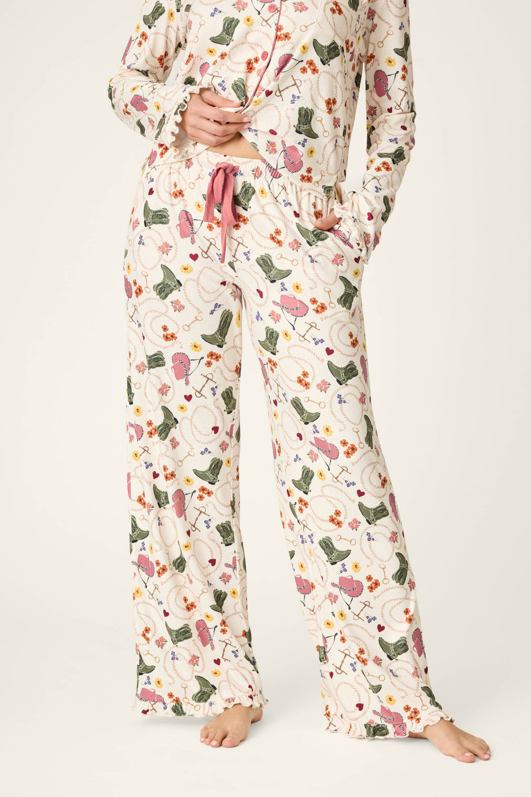 A person is wearing the P.J. Salvage Saddle Up Western Print Pajama Pants, featuring a pink drawstring and decorated with colorful floral and cactus patterns, while standing barefoot on a light background.