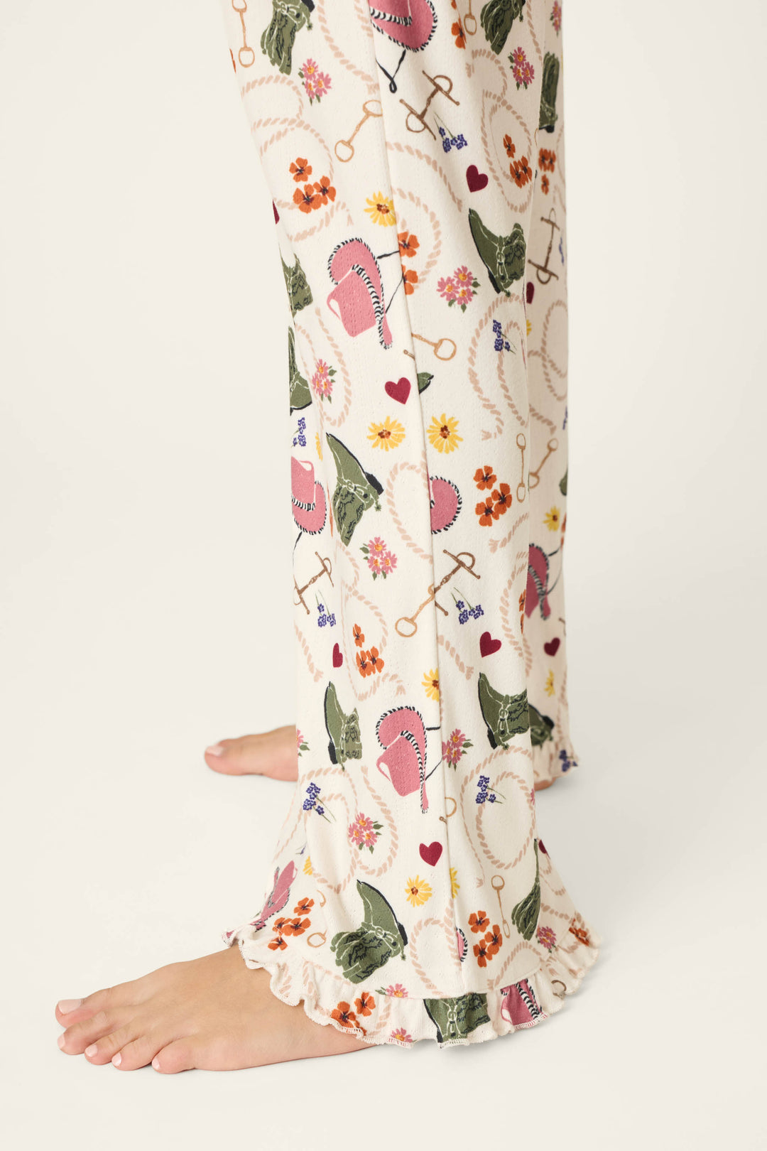A woman’s lower legs wearing the P.J. Salvage Saddle Up Western Print Pajama Pants, featuring a cream fabric with colorful floral, bird, and key designs and ruffled hems; bare feet are visible against a light background.
