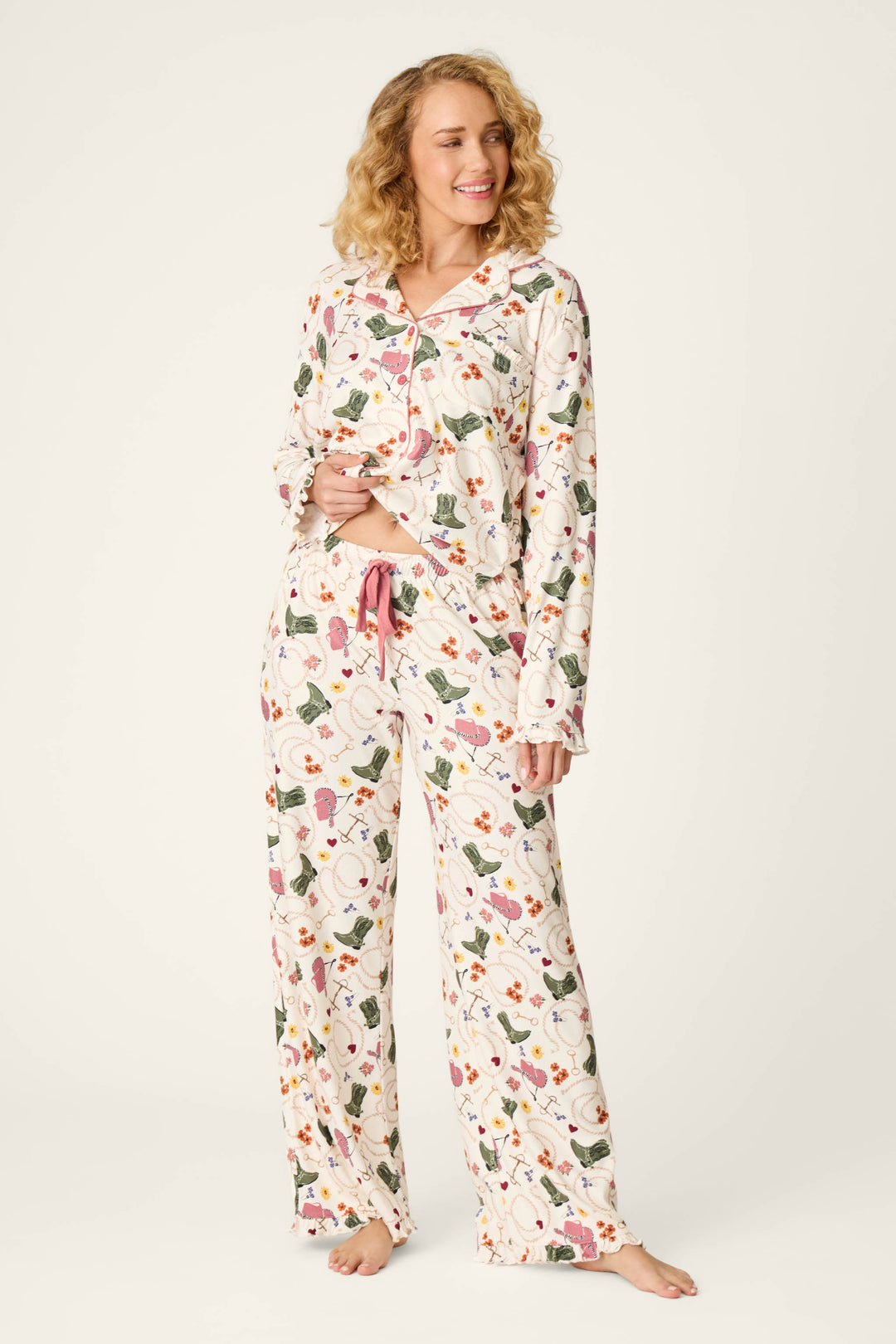 A woman with curly blonde hair stands barefoot and smiles against a plain light background, wearing the P.J. Salvage Saddle Up Western Print Pajama Pants. The pants feature a long, relaxed fit with button-up detailing, floral prints accented by pink trim and a pink drawstring at the waist.