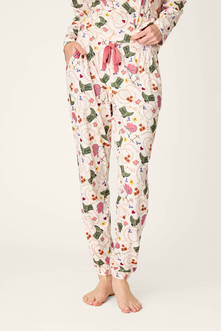 A person wearing the P.J. Salvage Saddle Up Western Print Pajama Jogger Pants, featuring a light-colored base with a vibrant floral and bird pattern, pink drawstring, and pockets. They are barefoot with hands in pockets, standing against a plain background.