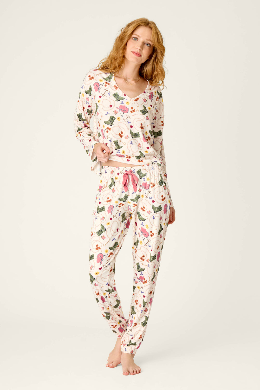 A woman with wavy red hair is standing barefoot against a plain light background, gently smiling at the camera while wearing P.J. Salvage’s Saddle Up Western Print Pajama Jogger Pants and a colorful long-sleeve top.