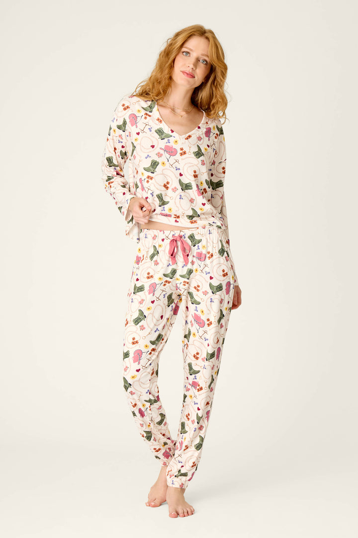 A woman with wavy red hair is standing barefoot against a plain light background, gently smiling at the camera while wearing P.J. Salvage’s Saddle Up Western Print Pajama Jogger Pants and a colorful long-sleeve top.