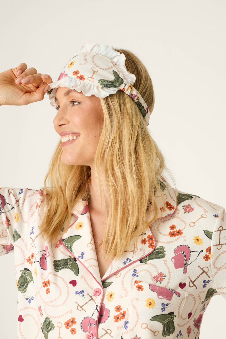 A woman smiles while wearing the P.J. Salvage Saddle Up Western Print 3-Piece PJ Set, complete with a matching ruffled sleep mask on her head, set against a light background.