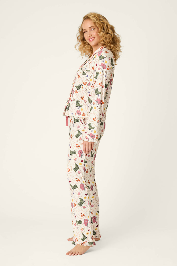 A woman with curly blond hair smiles and stands sideways, barefoot, wearing the P.J. Salvage Saddle Up Western Print Long Sleeve PJ Set on a plain background.