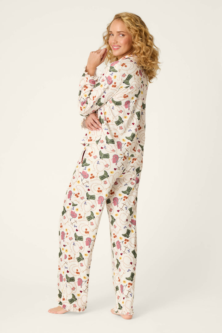 A woman with curly blonde hair wears the P.J. Salvage Saddle Up Western Print Long Sleeve PJ Set, standing barefoot on a white background, smiling and glancing over her shoulder with her hand touching her face.