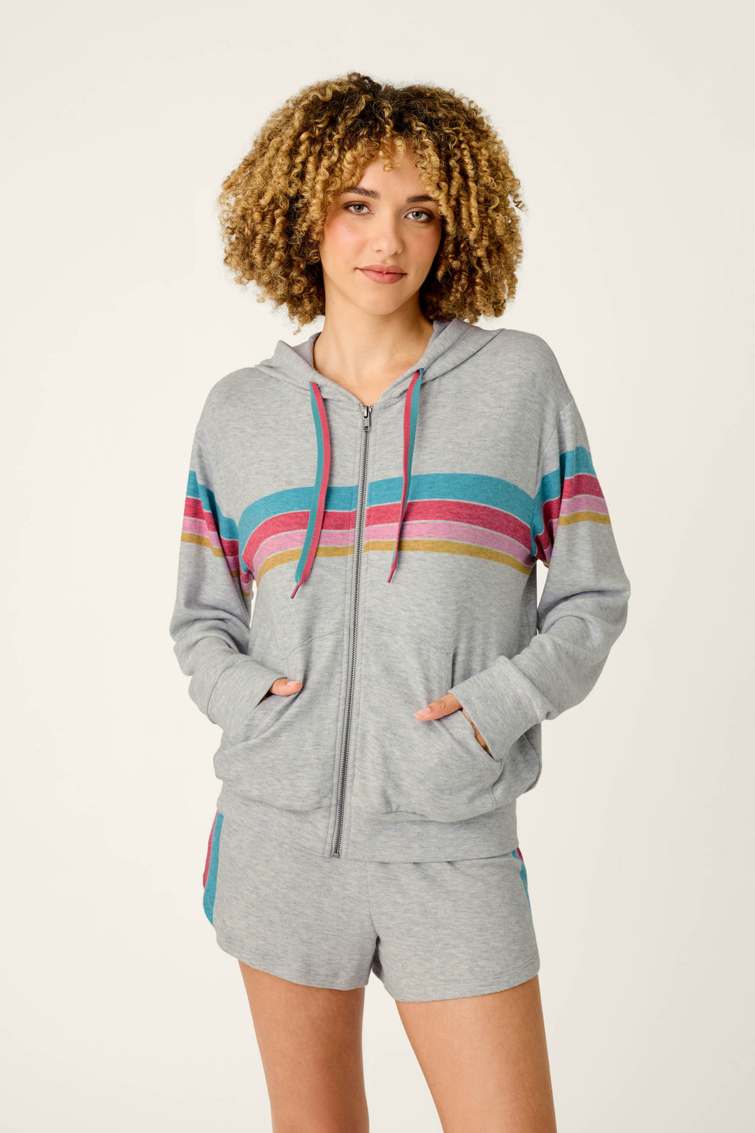 A person wearing the P.J. Salvage Takin’ It Easy Club Striped Zip-Up Hoodie and matching shorts with colorful horizontal stripes, standing with hands in pockets against a plain background.