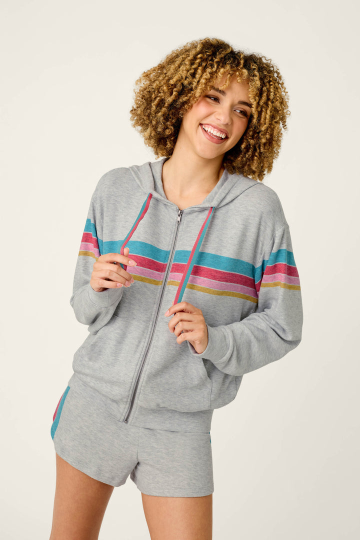 A smiling woman with curly hair wears the Takin’ It Easy Club Striped Zip-Up Hoodie by P.J. Salvage in light gray, featuring colorful stripes across the chest, paired with matching shorts and standing against a plain light background.