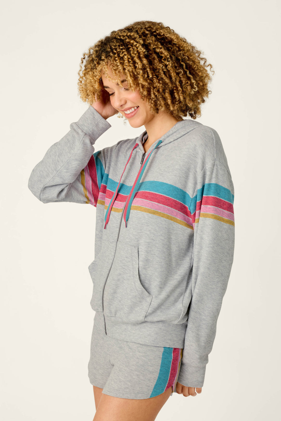 A woman with curly hair smiles while wearing the P.J. Salvage Takin’ It Easy Club Striped Zip-Up Hoodie and matching shorts featuring blue, pink, and yellow stripes across the chest and thigh. She touches her hair as she stands against a plain background.