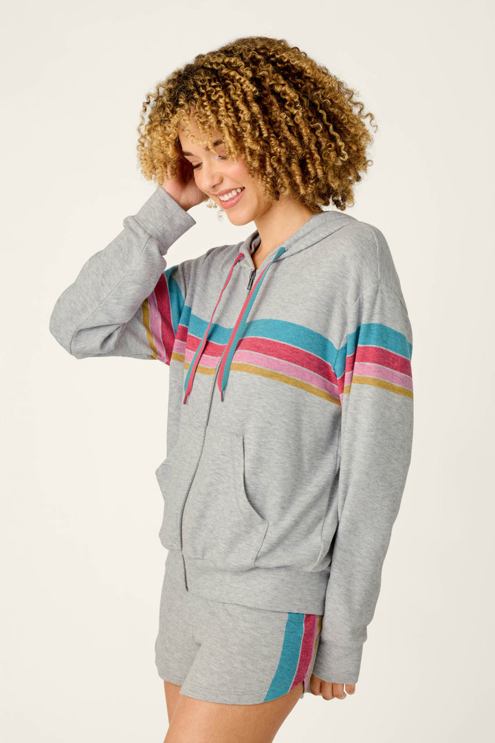A woman with curly hair smiles while wearing the P.J. Salvage Takin’ It Easy Club Striped Zip-Up Hoodie and matching shorts featuring blue, pink, and yellow stripes across the chest and thigh. She touches her hair as she stands against a plain background.