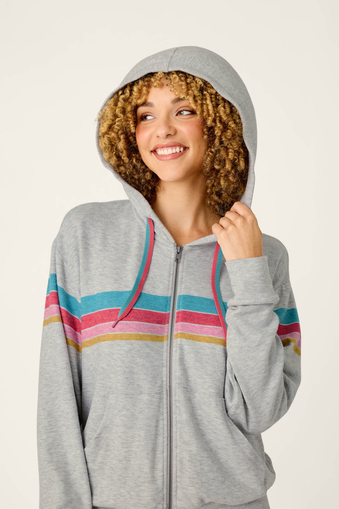 A smiling person with curly hair wears the P.J. Salvage Takin’ It Easy Club Striped Zip-Up Hoodie, featuring pink, blue, and yellow stripes and the hood up, holding the drawstring with one hand.
