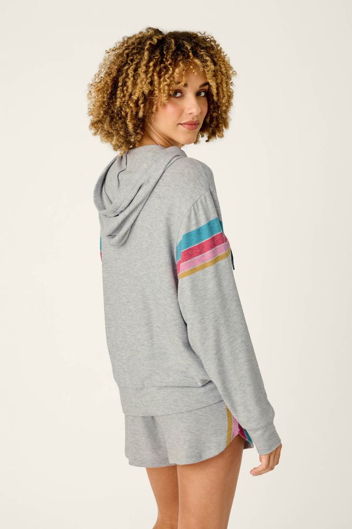 A person with curly hair is wearing the P.J. Salvage Takin’ It Easy Club Striped Zip-Up Hoodie and matching shorts, featuring colorful stripes on the sleeves and shorts, standing and looking back over their shoulder against a plain background.