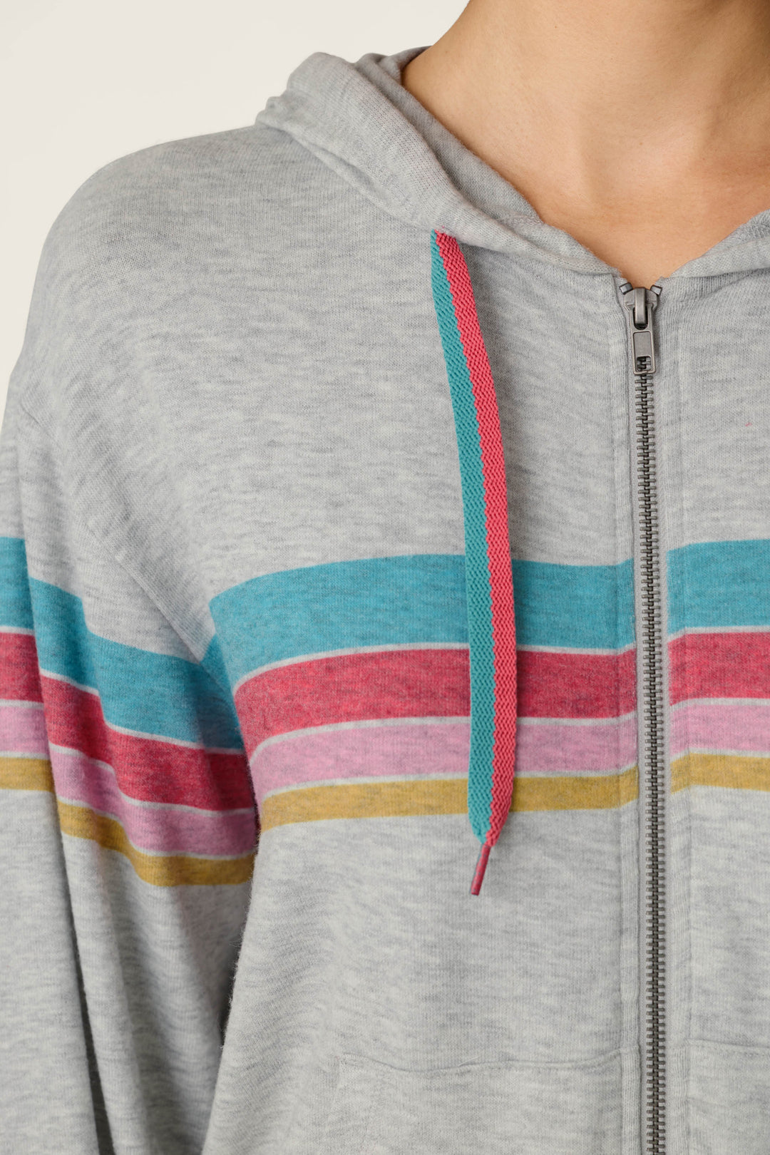A close-up of the P.J. Salvage Takin’ It Easy Club Striped Zip-Up Hoodie, featuring colorful horizontal stripes and mismatched teal and pink drawstrings, with the wearer’s head partially out of frame.