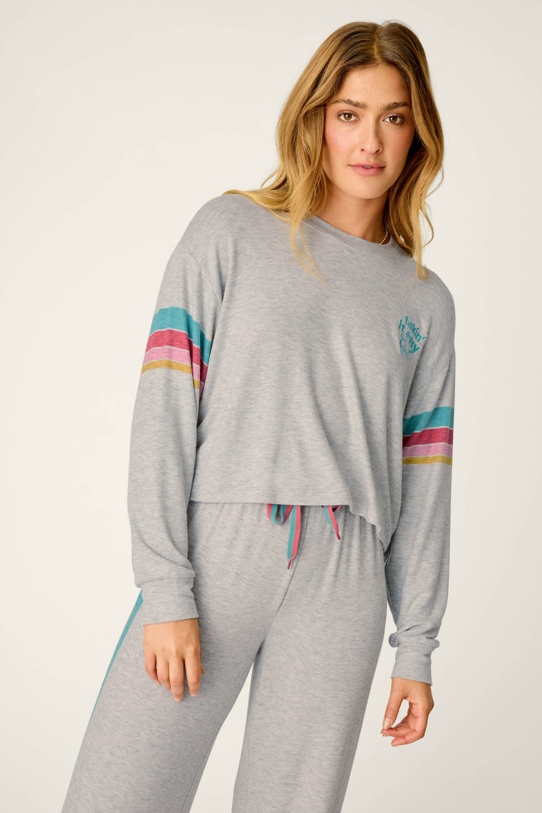 A woman poses against a neutral background wearing the P.J. Salvage Takin’ It Easy Club Striped Long Sleeve Top, a light gray lounge piece featuring colorful stripes on the arms.