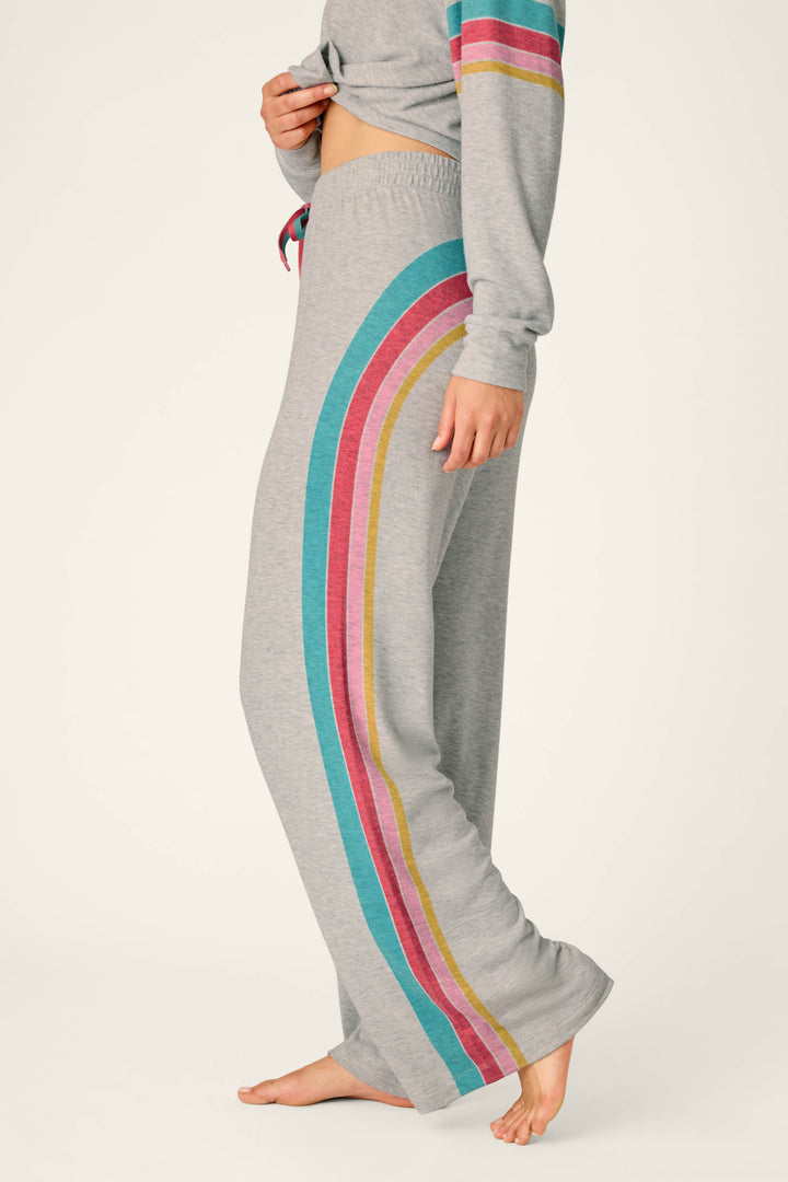 A person is standing barefoot on a light background, wearing the P.J. Salvage Takin’ It Easy Club Striped Lounge Pants, which feature gray fabric with teal, pink, red, and yellow stripes down the side.