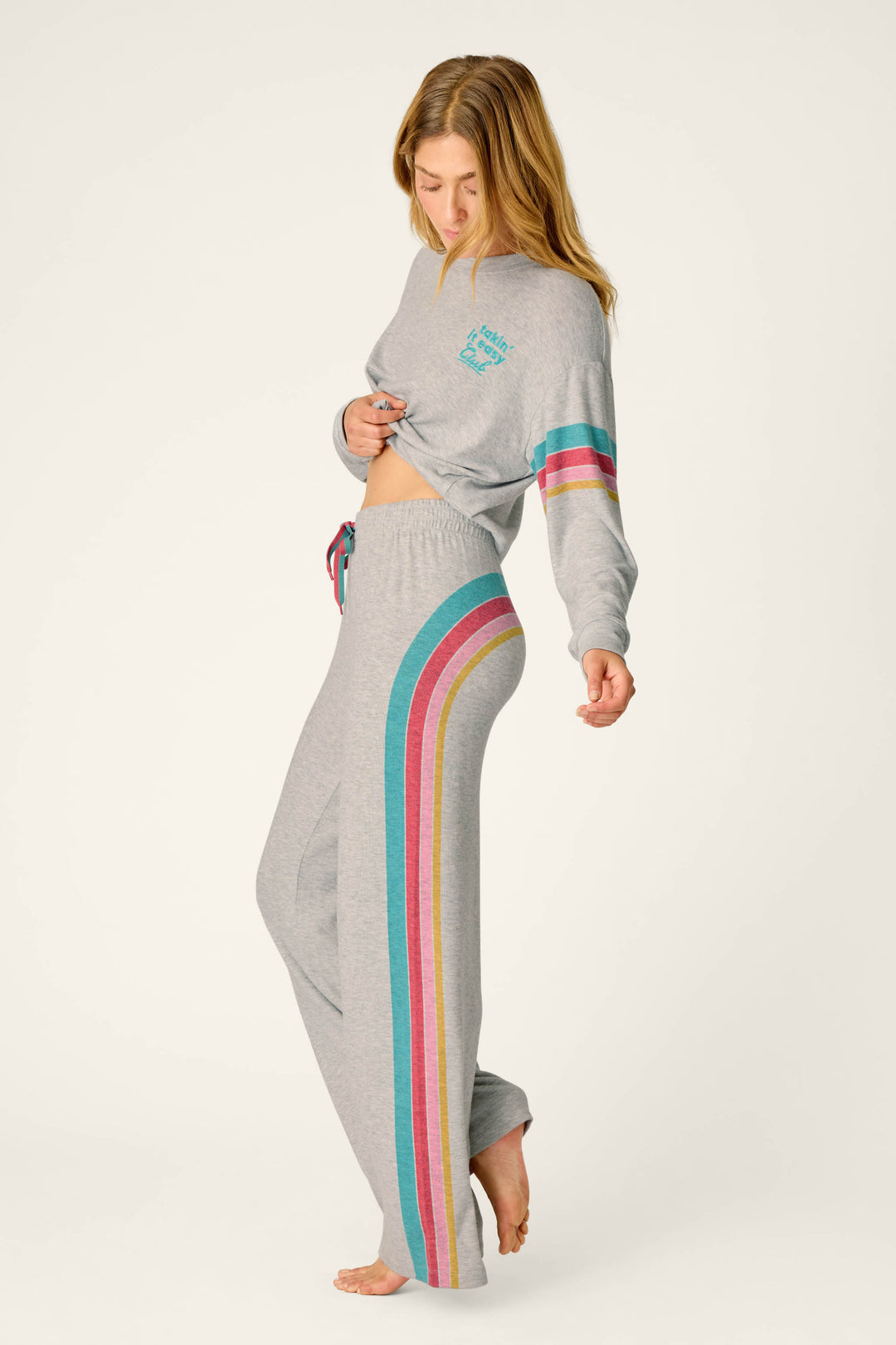 A woman stands barefoot on a plain background, slightly lifting her sweatshirt to reveal her midriff while wearing the P.J. Salvage Takin’ It Easy Club Striped Lounge Pants—a light gray loungewear set featuring colorful stripes down the side of the pants and sleeves.