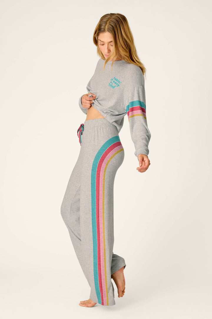 A woman stands barefoot on a plain background, slightly lifting her sweatshirt to reveal her midriff while wearing the P.J. Salvage Takin’ It Easy Club Striped Lounge Pants—a light gray loungewear set featuring colorful stripes down the side of the pants and sleeves.
