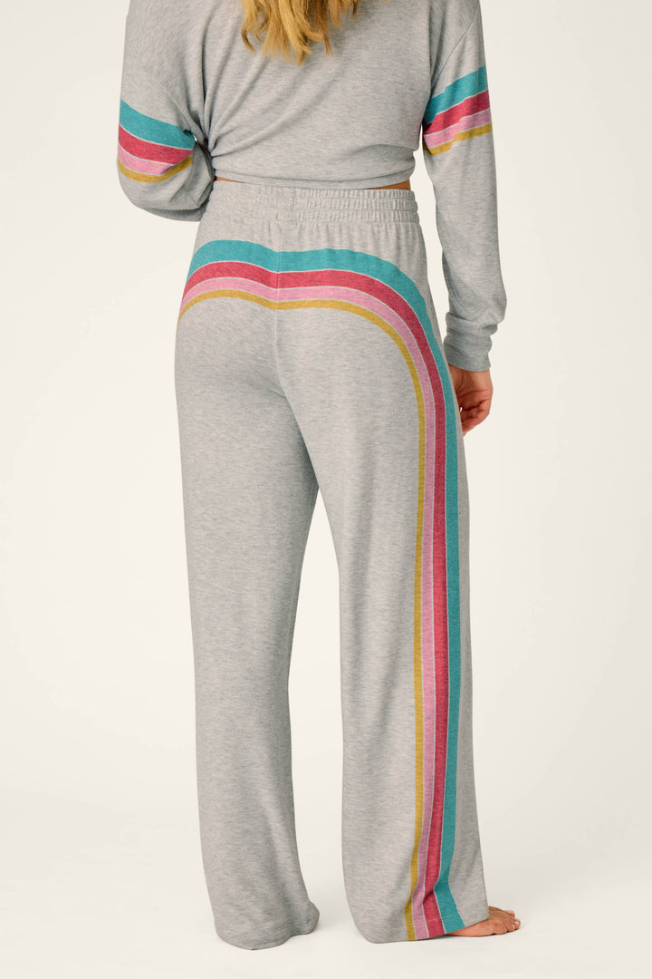 A person is wearing the P.J. Salvage Takin’ It Easy Club Striped Lounge Pants, featuring gray fabric with pink, red, gold, and teal stripes running horizontally across the back and down the side of the left leg. A matching long-sleeve top with coordinating sleeve stripes is partially visible.
