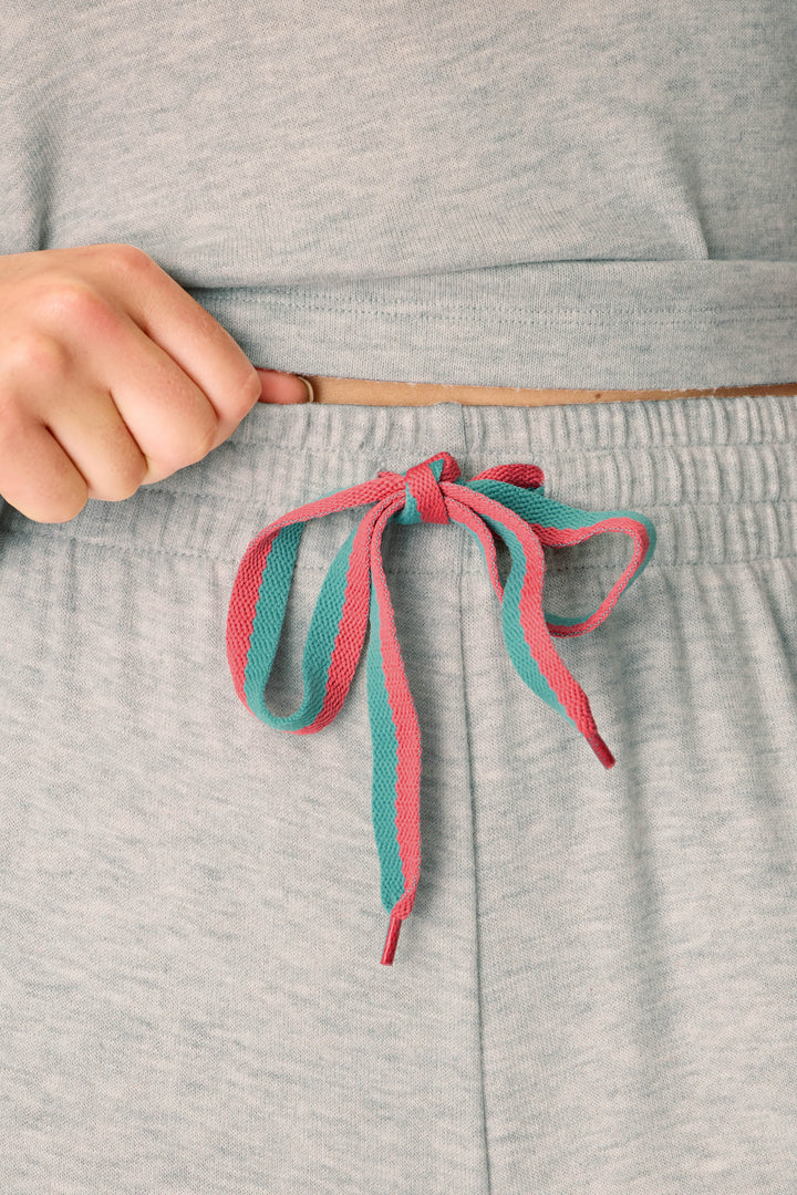 A close-up of a person’s hand adjusting the waistband of P.J. Salvage’s Takin’ It Easy Club Striped Lounge Pants, featuring a pink and teal drawstring tied in a bow.