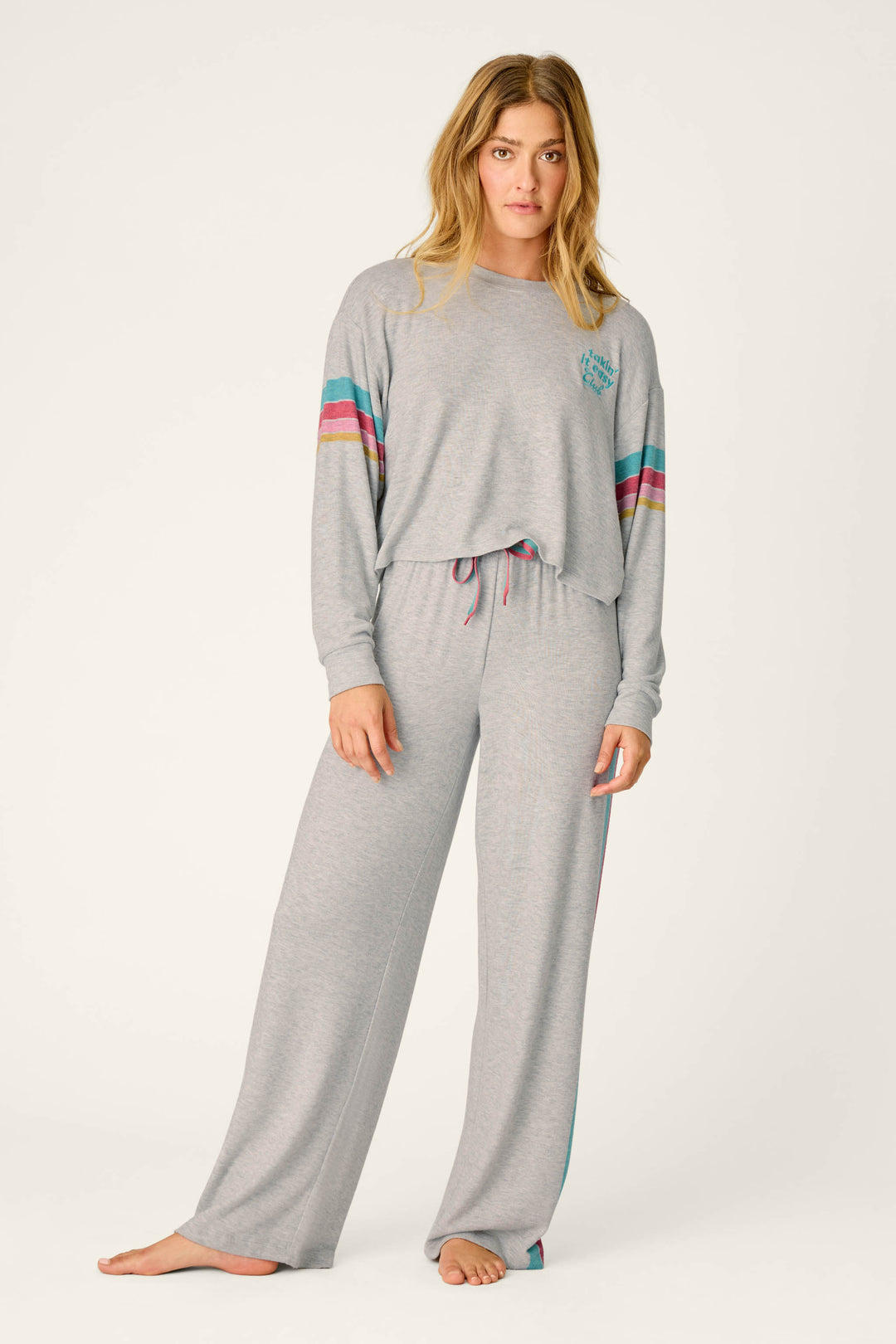 A woman stands barefoot, wearing the P.J. Salvage Takin’ It Easy Club Striped Lounge Set—a matching light gray ensemble with wide-leg pants and a long-sleeve top accented by colorful sleeve stripes. She faces forward against a plain, light background.