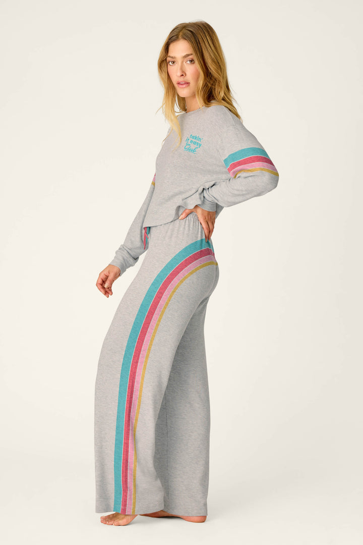 A woman stands in profile against a plain background wearing the P.J. Salvage Takin’ It Easy Club Striped Lounge Set, a grey ensemble featuring blue, pink, and yellow stripes down the sleeves and pant legs.