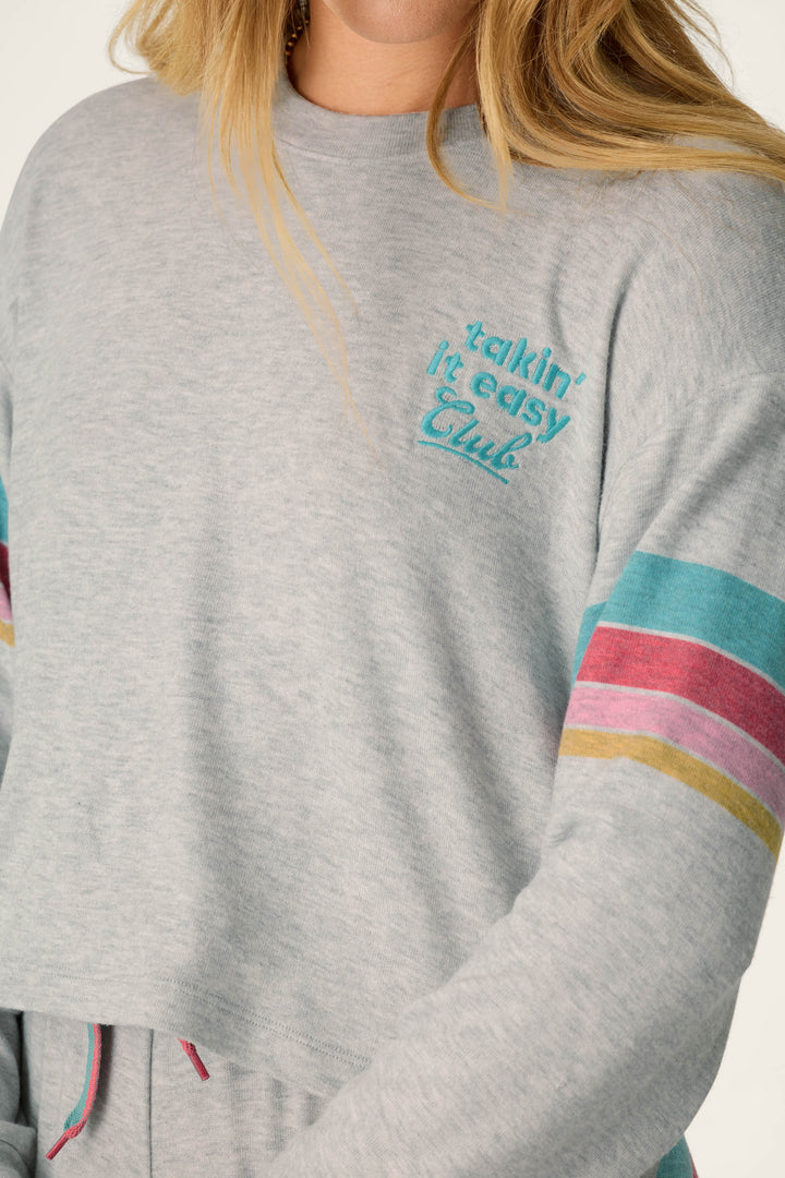 A person wearing the P.J. Salvage Takin’ It Easy Club Striped Lounge Set, featuring a light gray top with colorful sleeve stripes and “Takin’ It Easy Club” embroidered in blue on the chest. Their face is not fully visible.